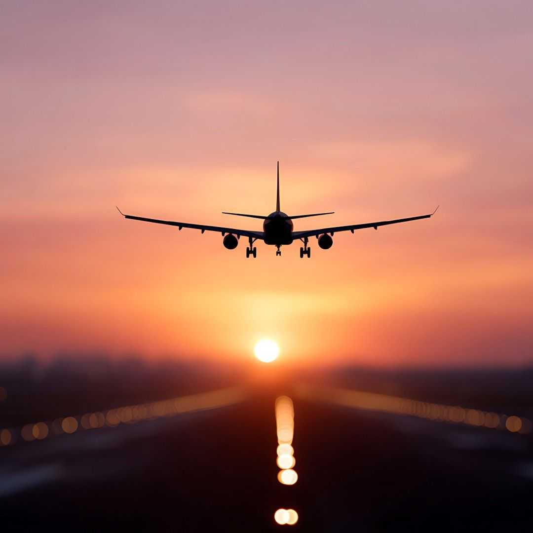 Airplane descending toward a runway at sunset, silhouetted against an orange sky with glowing runway lights leading toward the horizon.