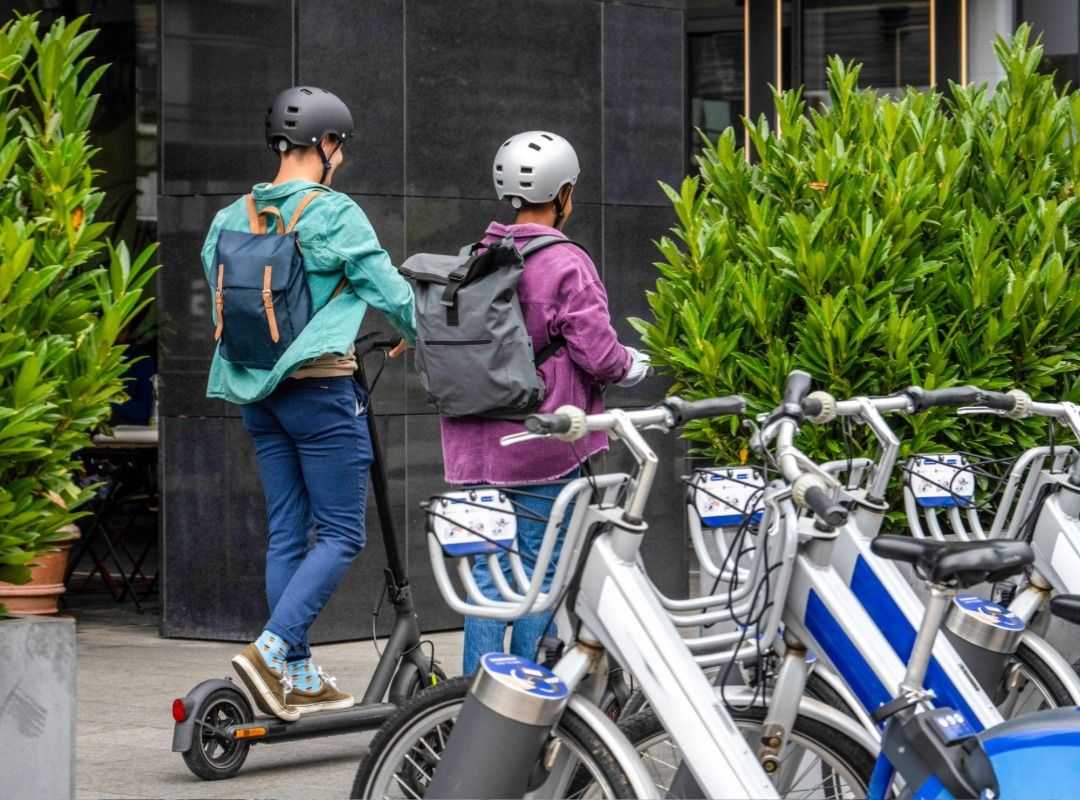 Two helmeted riders travel on an e-scooter beside a row of shared bicycles in an urban area with greenery and modern buildings in the background.